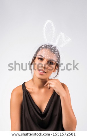 Cropped upper body's shot of a dark-haired girl, posing on a grey background. She is wearing black v-neck tank top and white headband with polka dot mesh rabbit ears and white polka dot mesh veil.