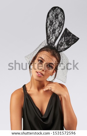 Cropped upper body's shot of a dark-haired girl, posing on a grey background. She is wearing black v-neck tank top and black headband with lace rabbit ears on her head and black mesh veil. 