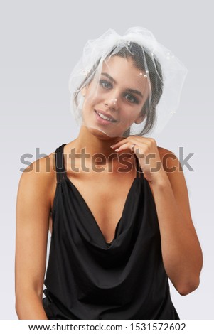 Cropped upper body's shot of a dark-haired girl, posing on a grey background. She is wearing black v-neck tank top and hairpin with short white mesh veil, decorating with beads. 