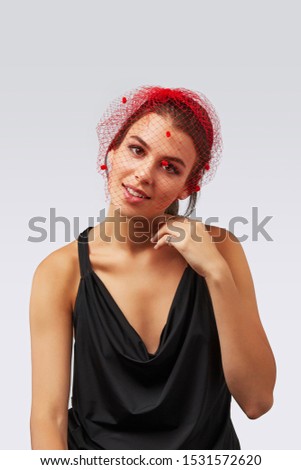 Cropped upper body's shot of a dark-haired girl, posing on a grey background. She is wearing black v-neck tank top and hairpin with red mesh veil, decorating with little red pompoms. 
