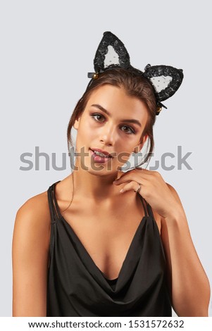 Cropped upper body's shot of a dark-haired girl, posing on a grey background. She is wearing black v-neck tank top and mesh kitten ears headband on her head, decorating with lace and bells. 