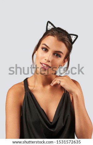 Cropped upper body's shot of a dark-haired girl, posing on a grey background. She is wearing black v-neck tank top and black polka dot mesh kitten ears headband on her head. Her left hand is near face
