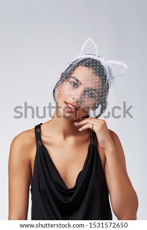 Cropped upper body's shot of a dark-haired girl, posing on a grey background. She is wearing black v-neck tank top and white polka dot mesh kitten ears headband on her head with polka dot mesh veil.