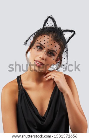 Cropped upper body's shot of a dark-haired girl, posing on a grey background. She is wearing black v-neck tank top and black polka dot mesh kitten ears headband on her head with polka dot mesh veil.