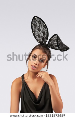 Cropped upper body's shot of a dark-haired girl, posing on a grey background. She is wearing black v-neck tank top and black headband with flower ornament lace rabbit ears on her head. 