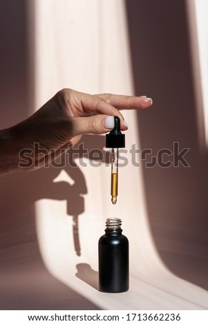 Cropped and vertical view of young adult woman holding pipette with natural oil over glass bottle, spill serum against pastel pink background with copy space
