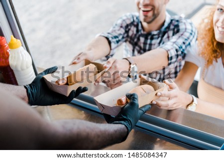 cropped view of african american man holding carton plates with hot dogs near happy customers 