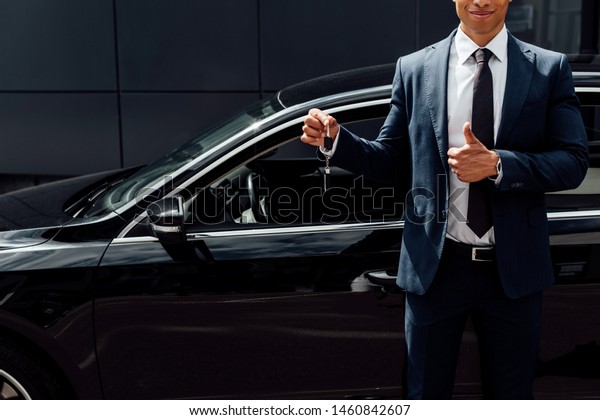 cropped view of african american\
man in suit holding car key  and showing thumb up near black\
car