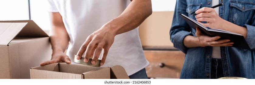 Cropped View Of African American Seller Putting Canned Food In Box Near Colleague With Notebook In Online Web Store, Banner