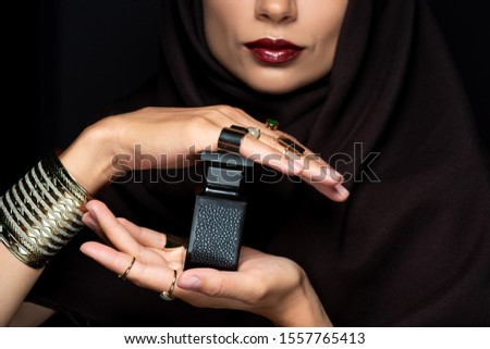 cropped view of beautiful Muslim woman in hijab with makeup in golden jewelry holding perfume isolated on black