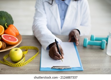 Cropped View Of Black Female Dietitian Planning Meal For Client, Writing In Clipboard, Sitting At Desk At Clinic. Weight Loss Specialist Writing Prescription For Patient At Medical Office