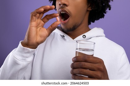 Cropped View Of Black Teenager Taking Pill, Holding Glass Of Fresh Water On Violet Studio Background, Panorama. Unrecognizable African American Adolescent Drinking Medication Or Vitamin