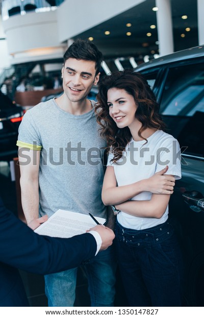 cropped view of car dealer holding clipboard with\
contract near happy woman and\
man