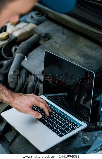 cropped view of car mechanic using laptop with blank\
screen near car 