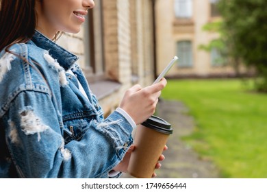 cropped view of cheerful student using smartphone and holding paper cup near university campus