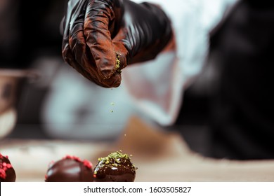 cropped view of chocolatier in black latex glove adding green pistachio powder on fresh made candies