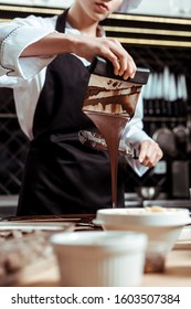 cropped view of chocolatier holding cake scraper with melted dark chocolate pouring on surface