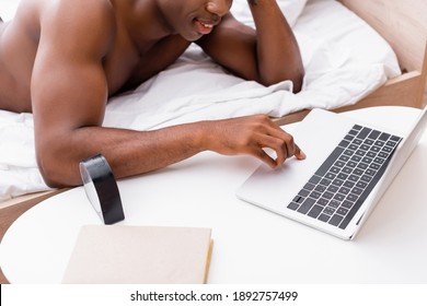 Cropped view of clock and book on table near smiling african american man using laptop on bed on blurred background - Powered by Shutterstock