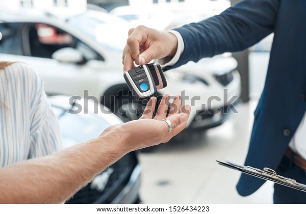 Cropped view of confident, professional salesman\
standing in auto salon and giving key into client hand. Concept of\
choosing new car in\
showroom