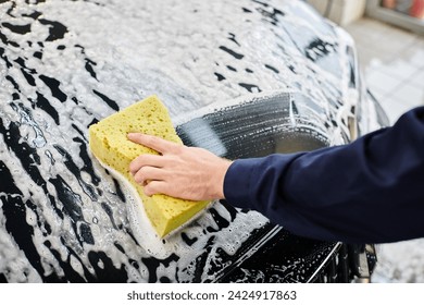 cropped view of devoted professional worker in blue uniform washing black car with soapy sponge