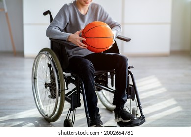 Cropped View Of Disabled Teenager In Wheelchair Holding Basketball At Home. Unrecognizable Handicapped Teen Boy Enjoying Active Lifestyle Indoors. Impairment And Sports Concept