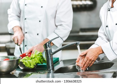 Cropped View Of Female Chef Washing Hands Over Sink While Colleague Cooking On Background In Restaurant Kitchen