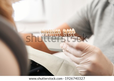Cropped view of the female dentist wearing white protective gloves matching teeth color of senior woman patient with palette in clinic. Teeth whitening concept