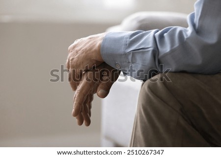 Cropped view, folded hands of unknown senior, man sitting on couch indoors. Close up shot of unrecognizable lonely retired male, deep in thoughts about senile diseases, loneliness in nursing house