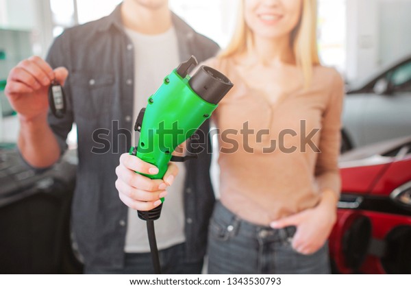 Cropped view of green\
electric charging plug in hand, electric car concept. Man holding\
car key. Young smiling family buying first electric car in the\
showroom. Green energy
