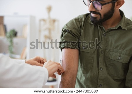 Cropped view of hand of general practitioner giving injection to boost immunity for bearded patient. Doctor in lab coat holding syringe with seasonal flu vaccine while young man sitting in exam room.