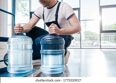 cropped view of happy delivery man holding blue bottles with water near hand truck