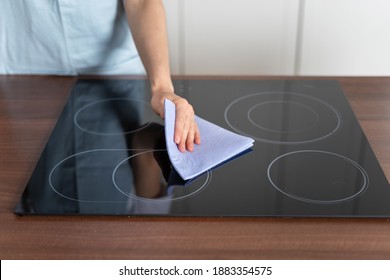 Cropped view of housewife washing electric stove with ceramic surface and wiping glass, using textile rag. Woman standing on modern kitchen at home