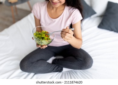 Cropped View Of Indian Lady Sitting Cross Legged On Bed At Home With Bowl Of Vegetable Salad, Eating Tasty Lunch. Unrecognizable Young Woman Enjoying Healthy Vegan Meal