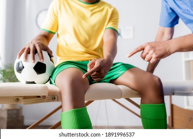 Cropped View Of Injured African American Football Player Pointing With Finger At Knee Near Doctor In Clinic