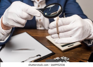 Cropped view of jewelry appraiser examining necklace with magnifying glass near calculator, clipboard and earnings on table isolated on grey
