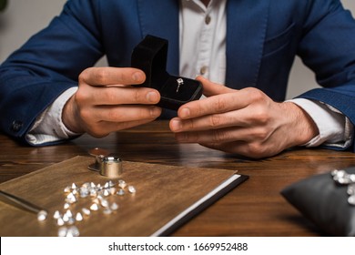 Cropped view of jewelry appraiser holding box with jewelry ring near gemstones on table on grey background