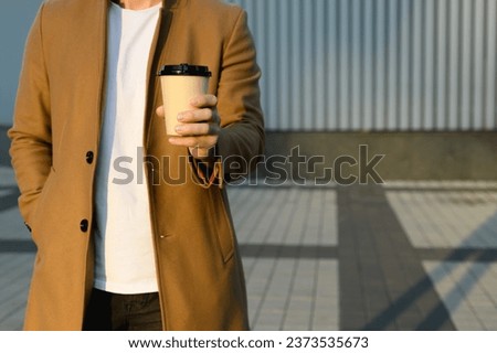 Cropped view of man in brown coat holding coffee to go and hand in pocket outdoors	