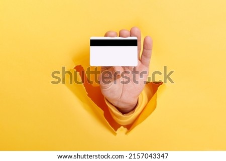 Cropped view of man hand breaks arm through paper and holding credit card, getting bank loan for shopping isolated over yellow background.