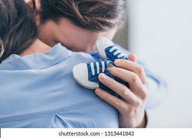 cropped view of man hugging woman and holding blue baby shoes in room, grieving disorder concept