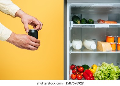 Cropped View Of Man Opening  Can With Soda Near Open Fridge With Fresh Food On Shelves Isolated On Yellow