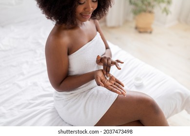 Cropped view of millennial Afro woman applying moisturizing hand cream, sitting on bed after shower at home. Young black lady pampering her skin after bath. Daily skincare routines concept