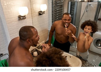 Cropped View And Mirror Reflection Of Black Adult Father And Little Son Brushing Teeth With Toothbrushes At Home Bathroom. Domestic Family Lifestyle. Fatherhood And Parenting. Morning Tome. Hygiene