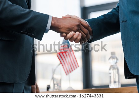 cropped view of multicultural diplomats shaking hands near flag of america