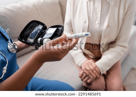 cropped view of multiracial caregiver holding lancet pen near senior woman sitting on sofa