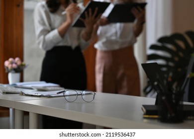 Cropped View Object Focused. A Glasses On A Table As A Foreground And A Two Business People Standing Behind As A Background In The Office. For Business And Work Concept.