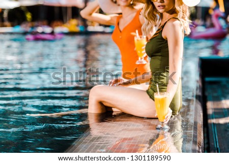 cropped view of pin up girls in swimsuits sitting at poolside with cocktails