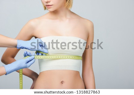 cropped view of plastic surgeon in latex gloves holding measuring tape on female breast with bandage, isolated on grey