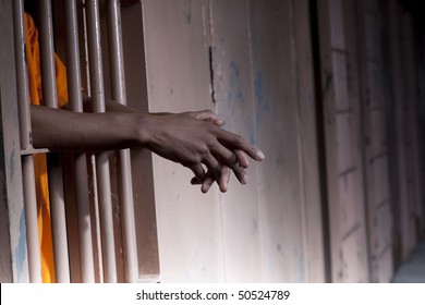 Cropped view of a prisoner in an orange jumpsuit standing in a prison cell with arms extended through the bars. Horizontal format.
