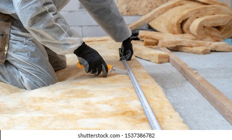 Cropped view of professional workman in protective workwear using knife and holding measuring tape over material, installing thermal insulation rock wool under the roof