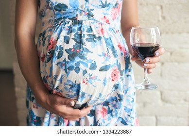 Cropped view, selective focus of glass of wine in hands of pregnant woman. Future mother embracing belly and drinking harmful alcohol while expecting baby. Concept of unhealthy lifestyle.